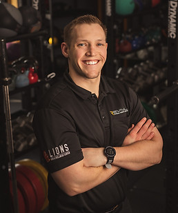 A confident male fitness trainer smiling in a gym, arms crossed, wearing a black polo shirt with logos and a watch, surrounded by exercise equipment.