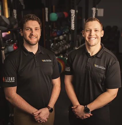 Two smiling men wearing black sports shirts with logos, standing confidently in a gym filled with fitness equipment.