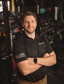 A man with curly hair smiling at the camera, arms crossed, wearing a black polo shirt with logos, standing in a gym filled with equipment.